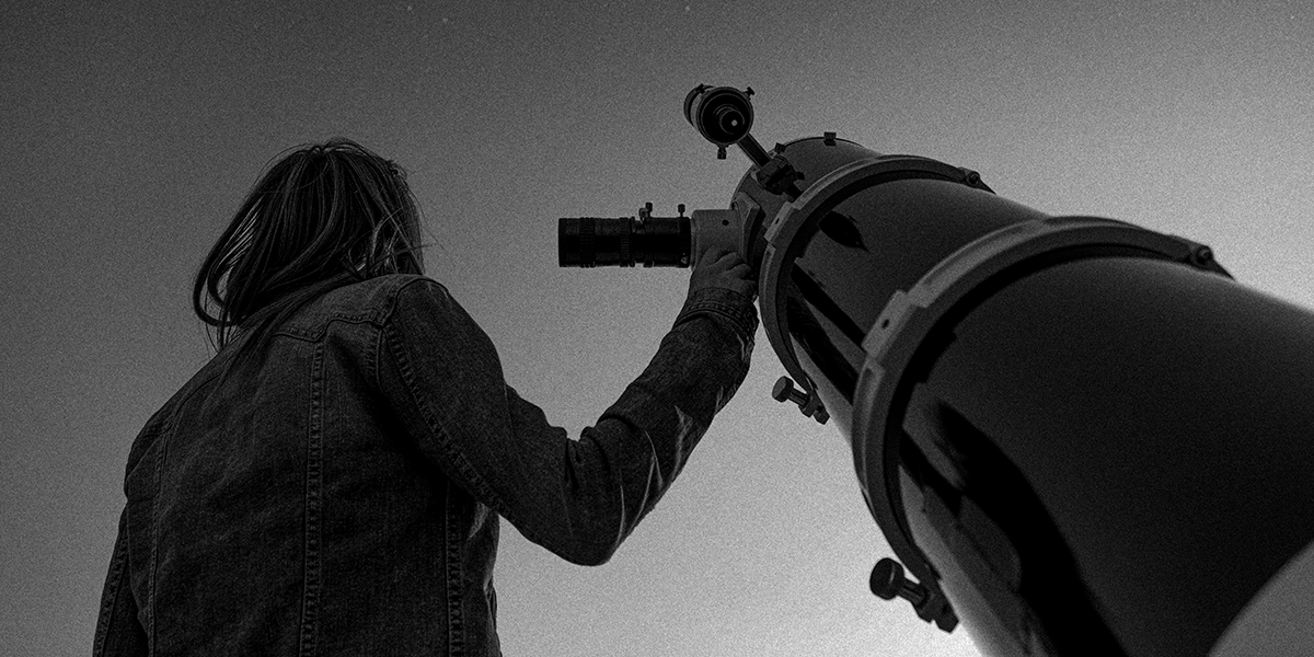 Black and white photo of person standing next to telescope and looking up at the sky