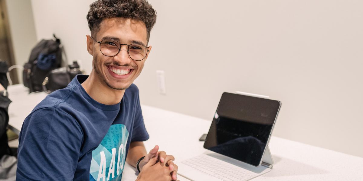 Student smiling at desk with tablet.